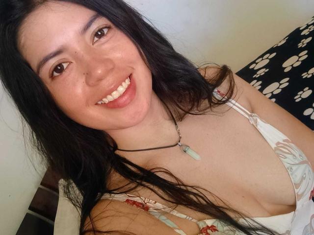 CristalDuport with long black hair, smiling, wearing a white floral dress, showing cleavage, necklace with green stone, brown eyes, fair skin, and visible freckles.