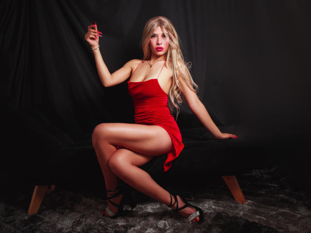 Martina Evans, met lang blond haar, een rode jurk en zwarte hakken, houdt een rode kaart vast, zit op een zwarte bank en laat haar benen en billen zien, met een sexy, zelfverzekerde blik..