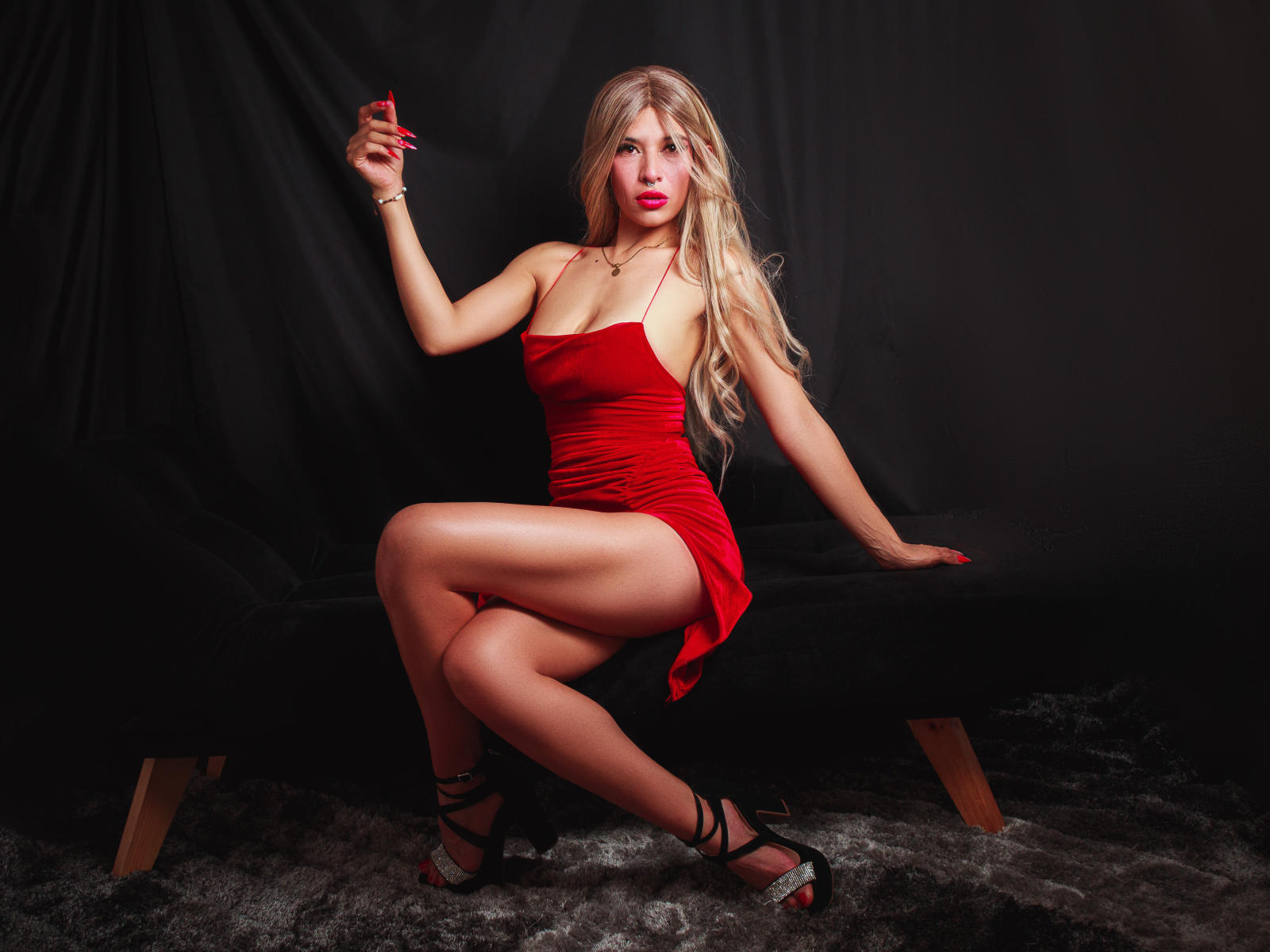 MartinaEvans with long blonde hair, red dress, and black heels, holding a red card, sitting on a black bench, showing her legs and ass, with a sexy, confident look.