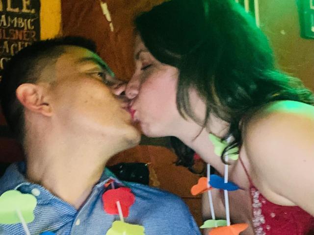 LucianSins is kissing LucianSins, both with eyes closed. LucianSins on left has short brown hair, blue shirt. LucianSins on right has long brown hair, red dress. Colorful fish sticks.