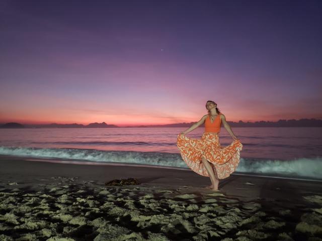 LucianSins, avec un haut orange et une jupe à motifs orange, tenant la jupe écartée, debout sur une plage rocheuse, coucher de soleil violet et rose, vagues de l'océan, ciel clair, peau légèrement humide, pieds nus..