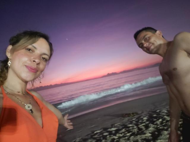 LucianSins, avec des cheveux bruns, une robe orange et des boucles d'oreilles, souriant à la caméra. LucianSins, torse nu, avec des cheveux foncés, regardant de côté. Coucher de soleil sur la plage, avec des vagues en arrière-plan..