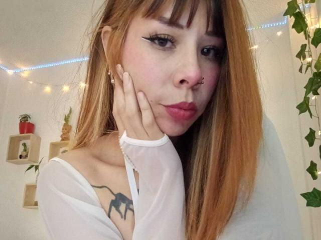 AriiannaHot with long brown hair, nose ring, and white top, showing off her spider tattoo on the right side of her neck, pouting with hand on cheek.