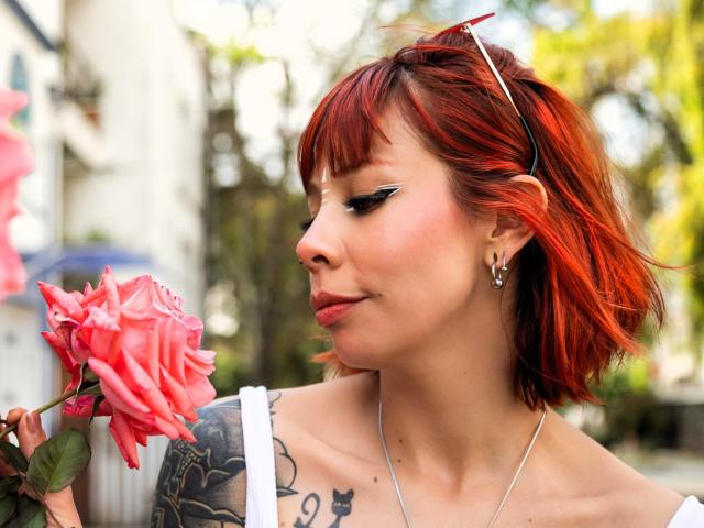 AriiannaHot with red hair, white dress, and tattoos, holds a pink rose, looking down. She's got black eyeliner, a nose piercing, and a silver necklace.