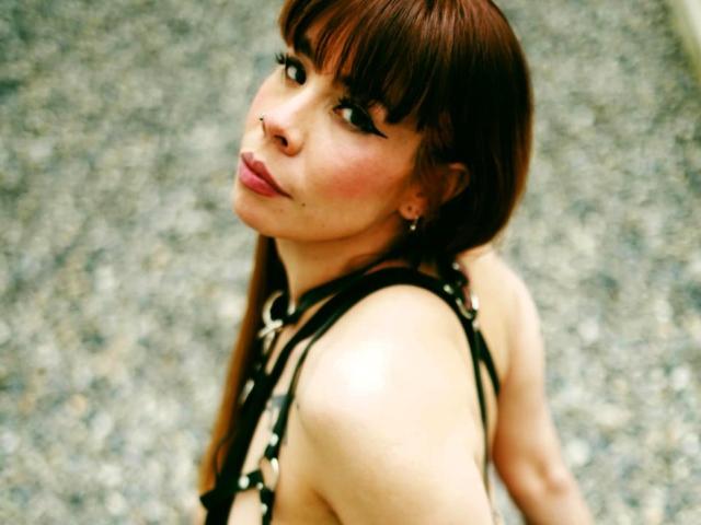 AriiannaHot with brown hair, bangs, and nose ring, wearing a black strappy top, looking back with pink lips, small earrings, and a subtle lip piercing.