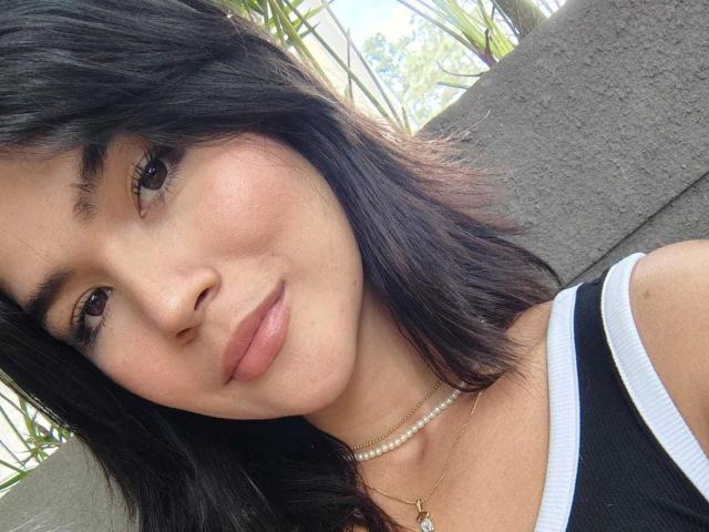 ScarlletZhen has medium-length black hair, brown eyes, and a subtle smile. She's wearing a black tank top with a white trim, a pearl necklace, and a small gold pendant.