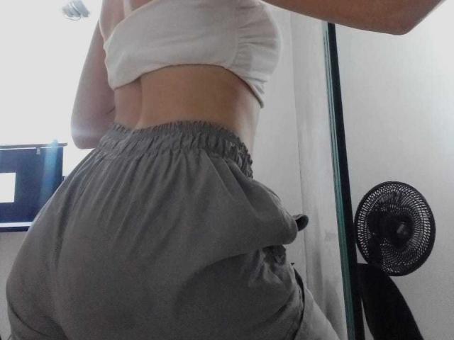 El trasero de MadisonAngel, con pantalones grises holgados y una camiseta blanca, está de pie con un abanico negro a la derecha y una caja azul en el fondo. Su espalda está hacia la cámara..