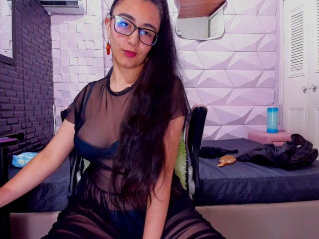 JewelryQueen, con cabello negro largo, gafas, aretes rojos y un vestido negro transparente que muestra sus senos, sentada en una silla en una habitación con una cama detrás de ella..