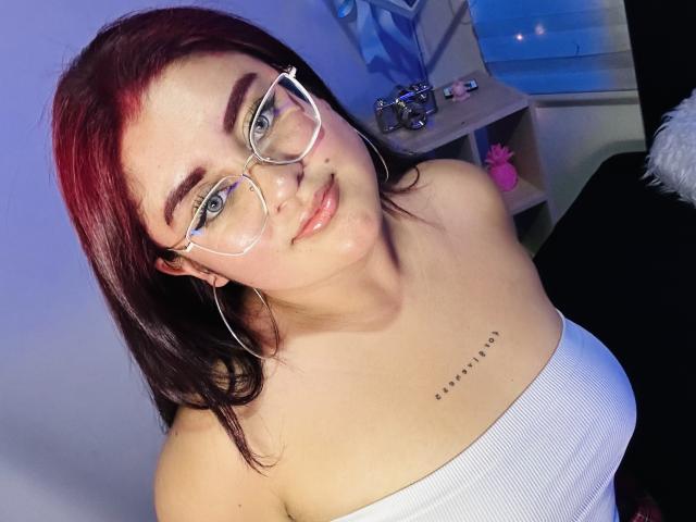 DaniielaaCorrtazaar with red-tinted hair, glasses, and white tube top, showing her tits and a small tattoo under her right breast. She has a shiny face and a cute smile.