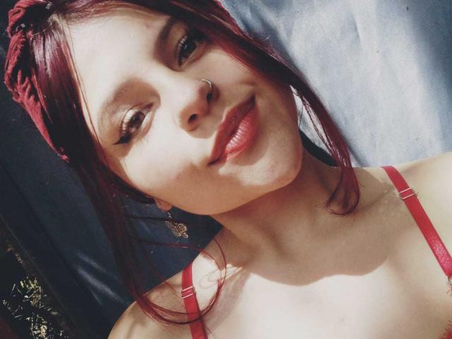 Sophie Hill ha trecce rosse, un piercing al naso e un reggiseno rosso, sorride con labbra lucide, mostrando il seno. Ha la pelle chiara, i capelli rossi e un trucco leggero..