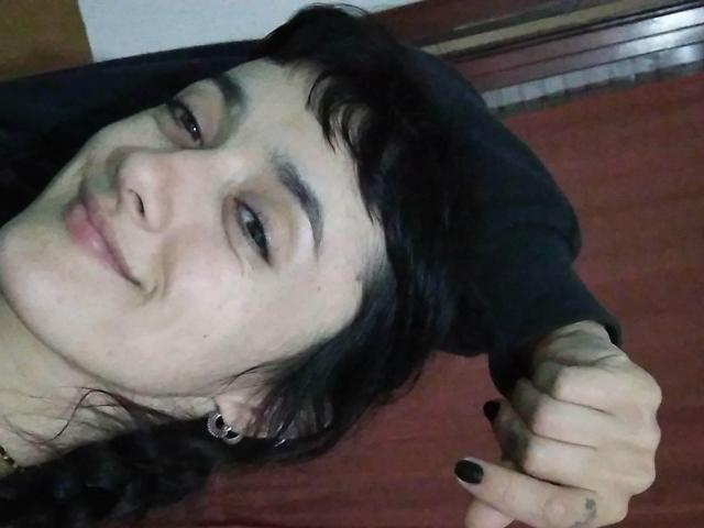 LaneLuce tem cabelo preto, um sorriso discreto e está piscando com um olho. Ela está segurando o cabelo com uma mão que mostra esmalte preto nas unhas e uma pequena tatuagem..