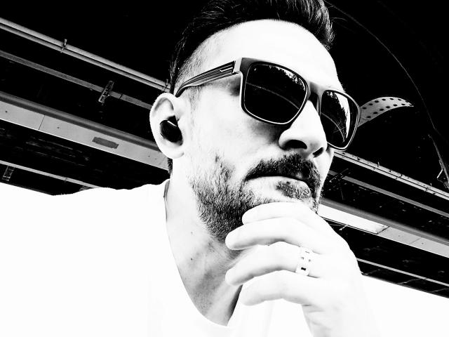 NeueHeimat-hot with a beard, sunglasses, and a ring on his finger, holding his chin with a white shirt, black and white image, looking serious, his hair is styled up.
