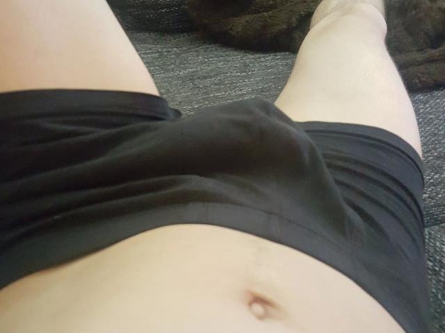 NeueHeimat-hot's black shorts are tight around their sizable, bulging cock. Their pale, flat stomach with a visible navel is in the foreground. Their legs are spread apart.