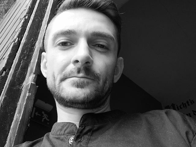 NeueHeimat-hot has a clean-shaven face, short dark hair, and a light beard, wearing a dark shirt, standing in a doorway, with a slight smirk. Black and white photo.