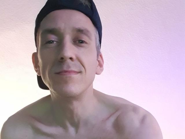 HarteLatte, sin camisa y con una ligera sonrisa, lleva puesto un gorro de béisbol azul oscuro, tiene una complexión delgada y musculosa, con clavículas visibles y un fondo rosado-morado sutil..