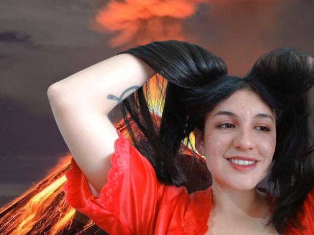 MargaritaDudole with black pigtails, smiling, wearing a red dress, has a tattoo on her left arm, and has visible freckles on her face. Her tits are small.