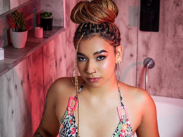 MadissonGreey with braided bun, nose ring, and hoop earrings, wearing a floral halter top, showing her tits, looking at the camera with a serious expression.