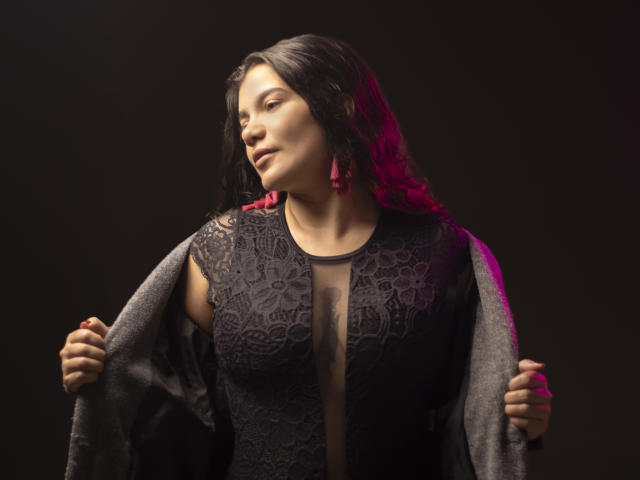 BulmaHot with dark wavy hair, wearing a black lace dress, holding a gray shawl, red earrings, eyes closed, slight smile, dark background, red light on right.