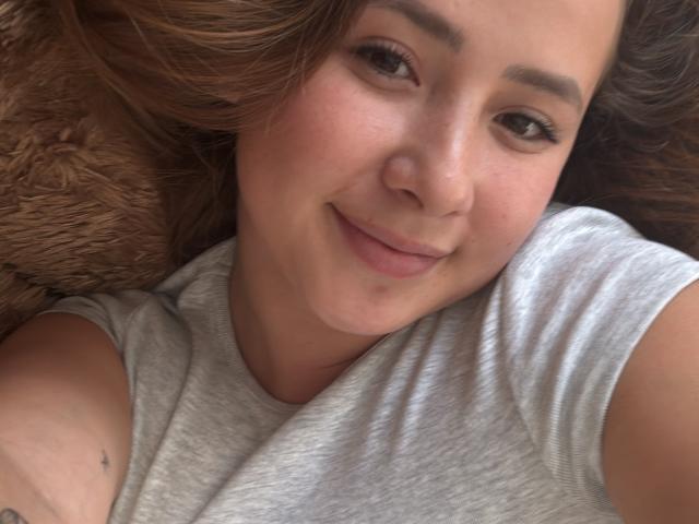 NatSorian's face with light brown hair, subtle smile, and brown eyes, wearing a gray shirt, lying on a brown furry surface, showing a small tattoo on her upper arm.
