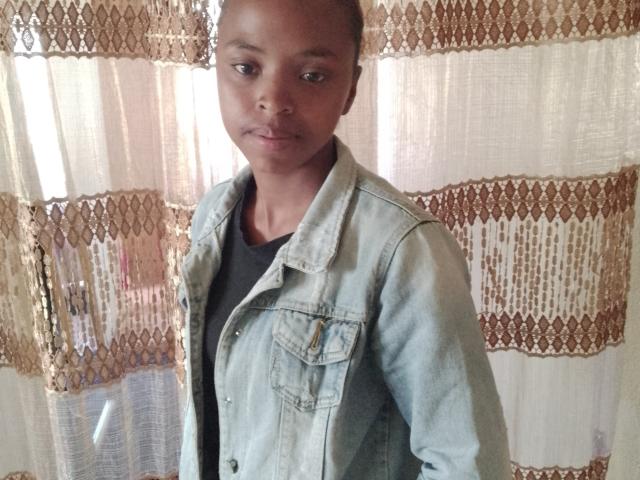 Nerina with dark skin, short hair, wearing a light blue denim jacket over a black shirt, standing in front of a curtain with brown and white patterns.