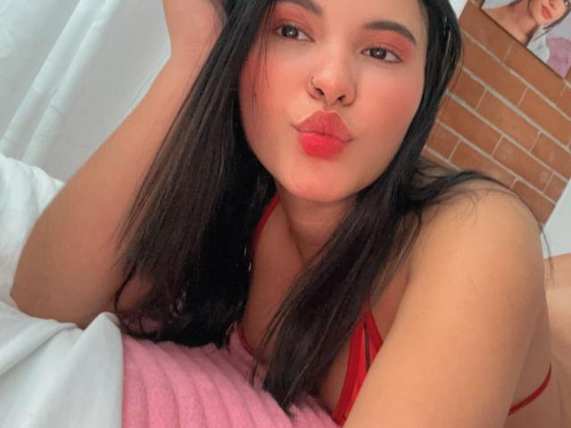 ArianaSmith69 with long black hair, red lipstick, and red bra, pouting her pink lips, lying on a pink blanket, showing her tits and ass. Brick wall in background.