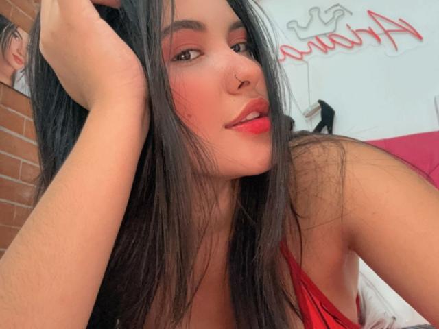 ArianaSmith69 with long black hair, red lipstick, and nose ring, touching her head, wearing a red top, showing her tits, in a selfie. Background is blurred.