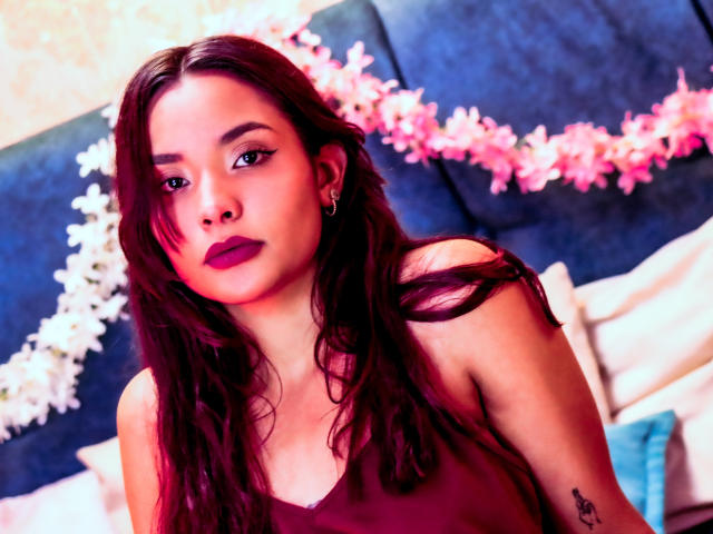 LeaKlarck has dark red lipstick, long brown hair, and a small tattoo on her right arm, wearing a burgundy top, looking at the camera with a serious expression.