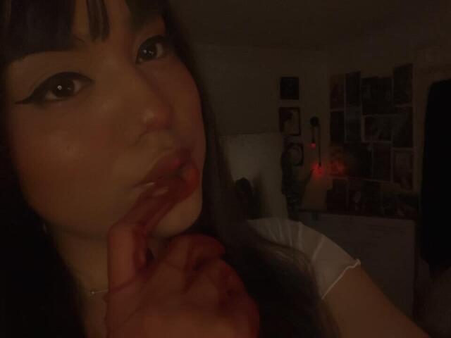SteffanyTaylor with dark eyeliner, biting her bloody finger in a dimly lit room, wearing a white shirt, her long black hair with bangs, red lipstick, and a necklace.