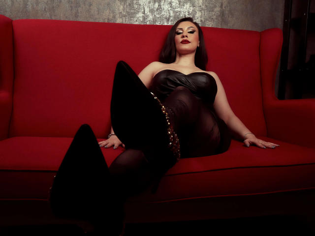 AmberCrost with pale skin, red lipstick, and black leather dress, lounging on red sofa, legs up, showing black stiletto boots with gold studs, confident, sexy.