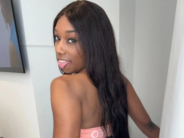 Niki-May-hot with long black hair, dark skin, and small tits, looking over her shoulder, tongue slightly out, wearing a pink top, in a bright indoor setting.