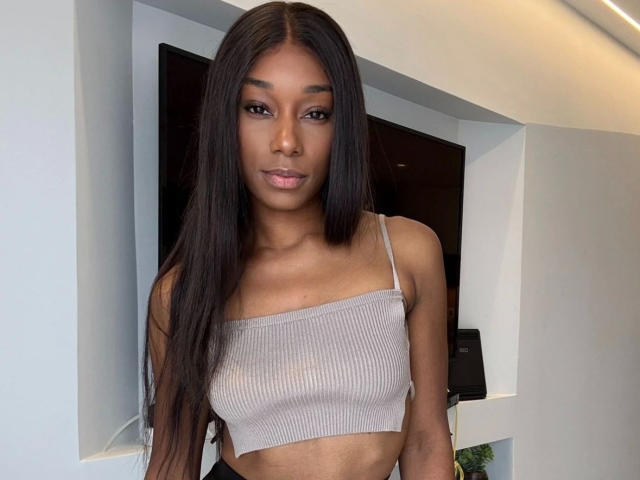 Niki-May-hot with long black hair, brown skin, and full lips wearing a beige ribbed crop top, showing her small tits. She's standing in a modern room.