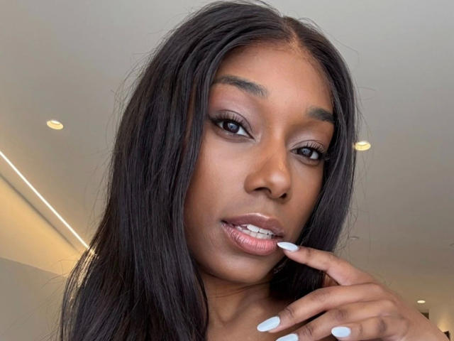 Niki-May-hot with long black hair, dark brown skin, and white nails, biting her finger with her shiny pink lips, looking at the camera with seductive, half-closed eyes.
