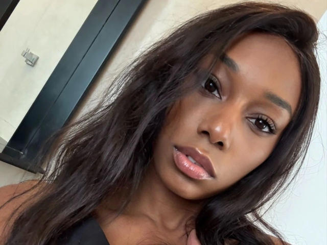Niki-May-hot has dark brown skin, long wavy black hair, and shiny pink lips with subtle makeup. She's looking directly at the camera with a slightly parted mouth.