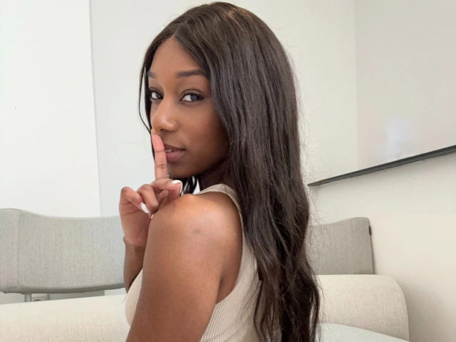 Niki-May-hot with long dark hair, brown eyes, and tan skin, wearing a beige tank top, holding a finger to her lips, looking over her shoulder. Boobs partially visible.