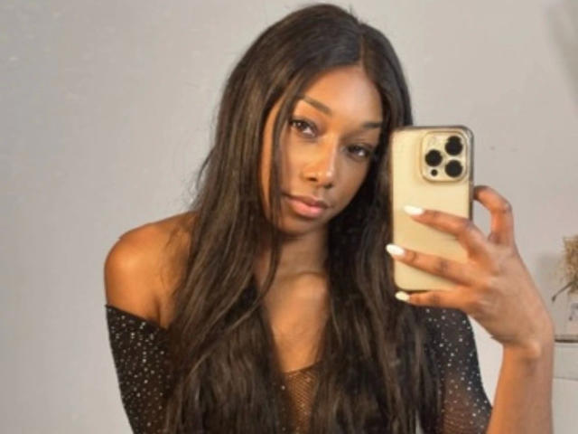 Niki-May-hot with long black hair, wearing a black sparkly off-shoulder top, holding a gold iPhone, looking at the camera, with white nails, and a serious expression.