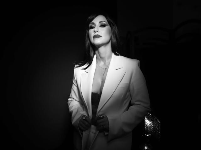 MissVonTease stands in dark, wearing a white blazer, exposing her tits in a black lace bra, with dark lipstick and shadowed eyes, holding a glittery purse. Black-and-white photo.