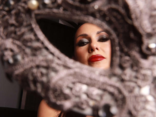 MissVonTease's face with red lipstick, dark eye makeup, and a confident expression, framed by a blurry, ornate, silver-gray mirror with small pearl-like details.