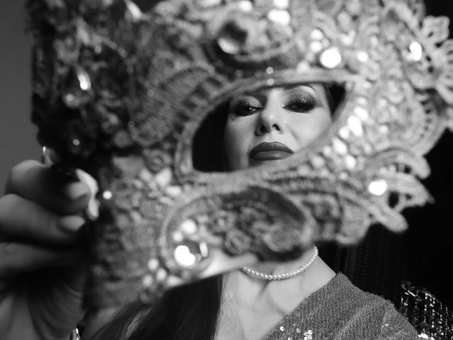 MissVonTease's face in a black-and-white photo, wearing a glittery, ornate hat with sparkles, dark makeup, and a pearl necklace, holding the hat in front of her.