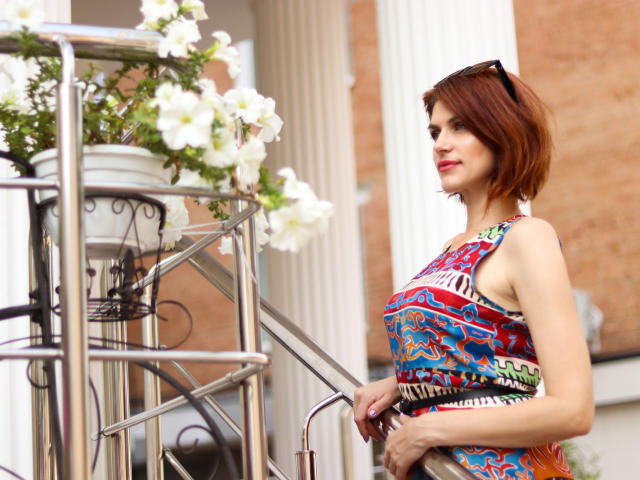 LisaTreds with red bob, wearing colorful, patterned dress, holding railing, white flowers in pot, black sunglasses on head, smiling, standing outside, holding metal staircase.