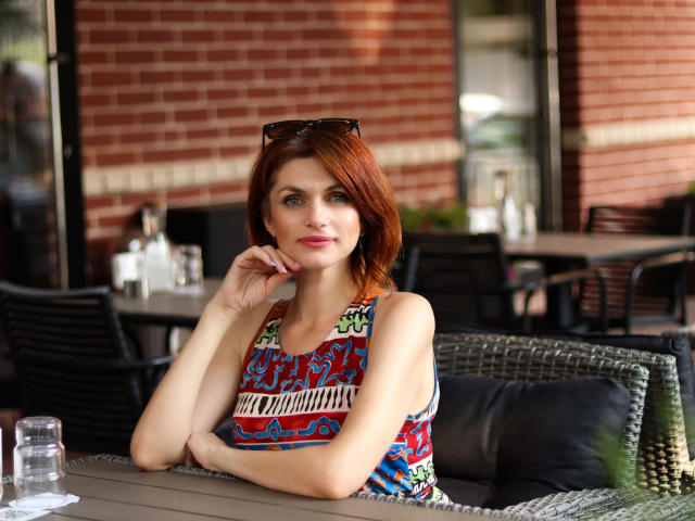 LisaTreds with red hair, blue eyes, and a colorful, patterned top, sits at an outdoor table, resting her chin on her hand, with sunglasses on her head.