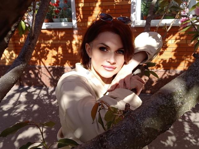 LisaTreds with red hair, blue eyes, and light skin, wearing a beige hoodie, gripping a tree branch, sunglasses on her head, sunlight casting shadows, smiling softly.