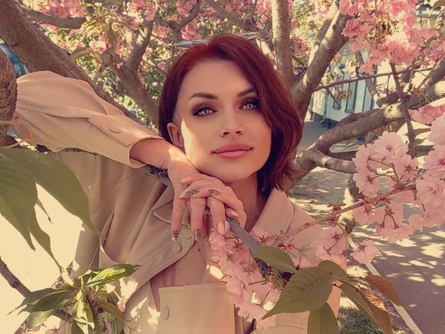 LisaTreds with pink blossoms, tan jacket, red hair, and pink nails, leaning on a tree branch, smiling with green eyes, sunlit, hands resting on her chin.