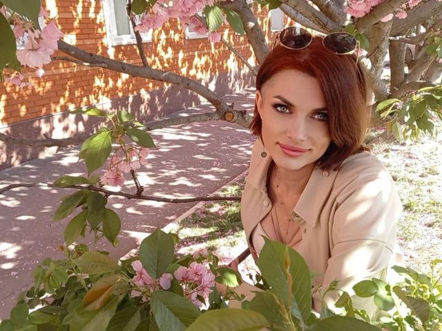 LisaTreds with red hair, blue eyes, and pink lipstick, wearing a beige jacket, standing in front of pink cherry blossoms, holding sunglasses in her hair.