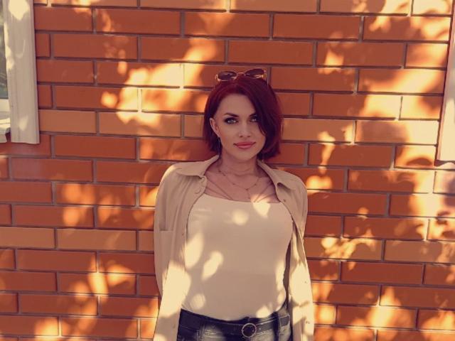 LisaTreds with red hair, wearing a beige shirt over a white top, black pants, and sunglasses on her head, standing against a brick wall with sun shadows.