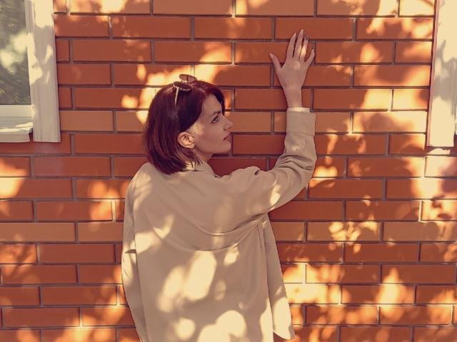 LisaTreds stands against an orange brick wall, wearing a beige coat, with her right hand touching the bricks, shadowed by tree branches. She has short brown hair, looking up.
