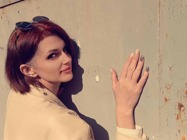 LisaTreds with short red hair, sunglasses on head, beige coat, side glance, smiling, showing hand with glittery nails, standing against a rough wall.
