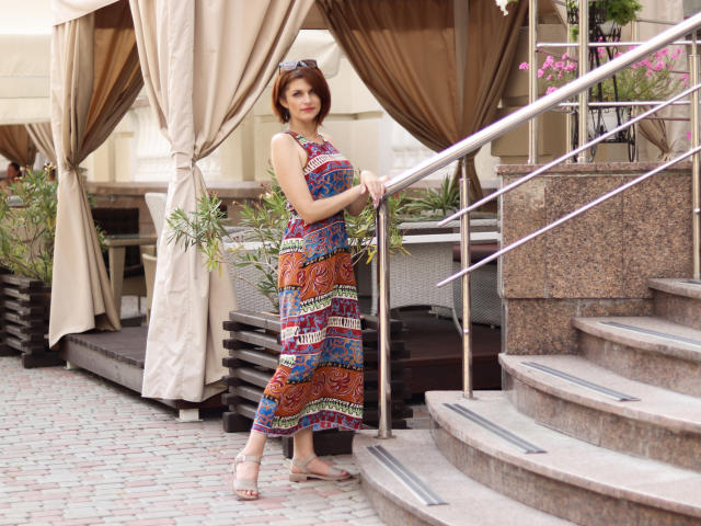 LisaTreds stands on a brick patio, holding a metal railing, wearing a colorful, patterned dress, and brown sandals, with her brown hair and sunglasses on her head.