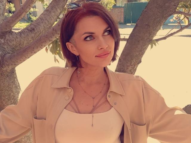 LisaTreds with short brown hair, wearing a beige jacket and white top, has a slim face, small tits, and a delicate necklace, standing under a tree in sunlight.