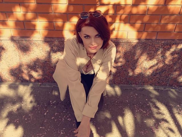 LisaTreds with red hair, wearing a beige jacket and black pants, crouching, looking up with blue eyes, sunglasses on head, shadowed brick wall, and sunlit pavement.