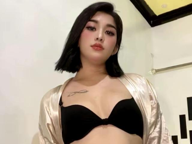 AsianBigCum with black bob, wearing shiny satin robe, black bra, showing small tits, tattoo on right chest, pink eyeshadow, and red lipstick, in dimly lit room.